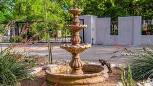 Una fuente de piedra en un jardín con un sendero empedrado y un muro blanco al fondo.
