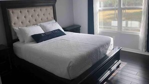 A bedroom with a large bed, a nightstand, a window with curtains, and a view of greenery outside. - Casa Padre, Perfect for long term stays. (Weslaco)