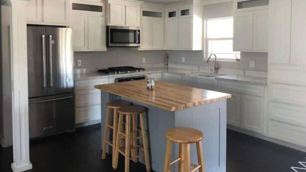 A modern kitchen with white cabinetry, stainless steel appliances, a central island with stools, and a window providing natural light. - Casa Padre, Perfect for long term stays. (Weslaco)