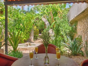 Une table avec deux verres de jus d’orange et un vase rempli de fleurs, le tout entouré d’une végétation luxuriante et d’un mur de pierre.