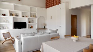 A modern living room with a white sofa, a dining table with a bowl of fruit, and a TV on a white cabinet. - Le Ciel de Marseille , T3 Lumineux Avec une vue Exceptionnelle (Marseille)