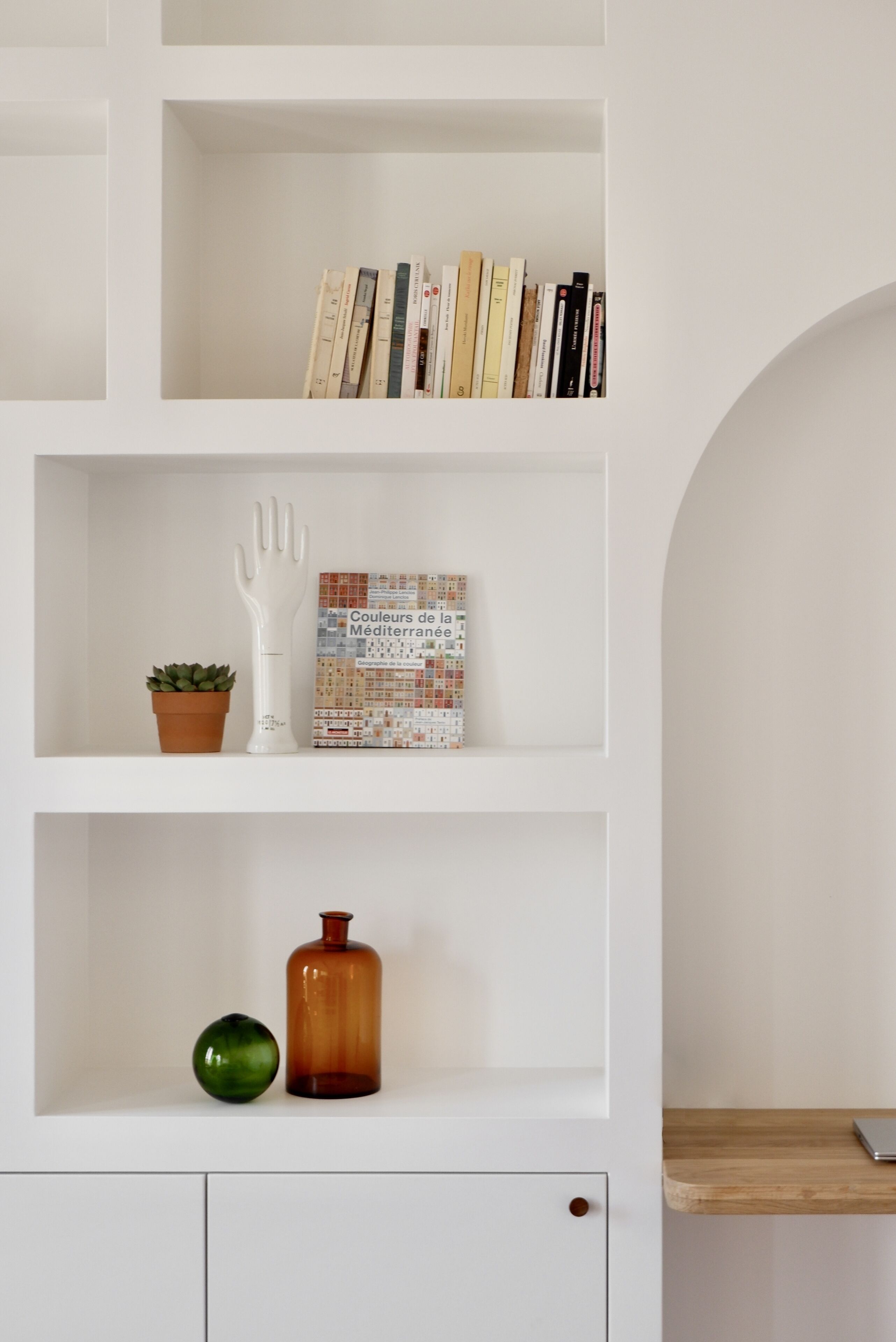 Ein weißes Regal mit Büchern, einem weißen, handgeformten Gegenstand, einer kleinen Topfpflanze und einer braunen Vase.