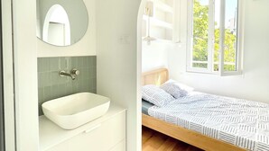 A modern bedroom with a built-in white sink, a bedside table, and a window with a view of greenery. - Le Ciel de Marseille , T3 Lumineux Avec une vue Exceptionnelle (Marseille)