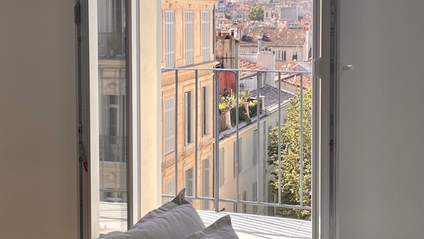 A room with a large window offering a view of a cityscape with historic buildings and a hill. - Le Ciel de Marseille , T3 Lumineux Avec une vue Exceptionnelle (Marseille)