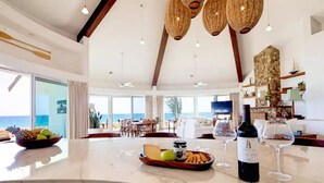 A spacious living area with a large window overlooking the ocean, a marble island with snacks and wine, and a modern interior design. - Luxury Beachfront Gem ~ Panoramic Sea Views ~ Heated Pool (B.C.S.)