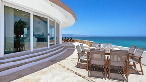 A seaside villa with a curved exterior, a dining area with a table and chairs, and a view of the ocean. - Luxury Beachfront Gem ~ Panoramic Sea Views ~ Heated Pool (B.C.S.)