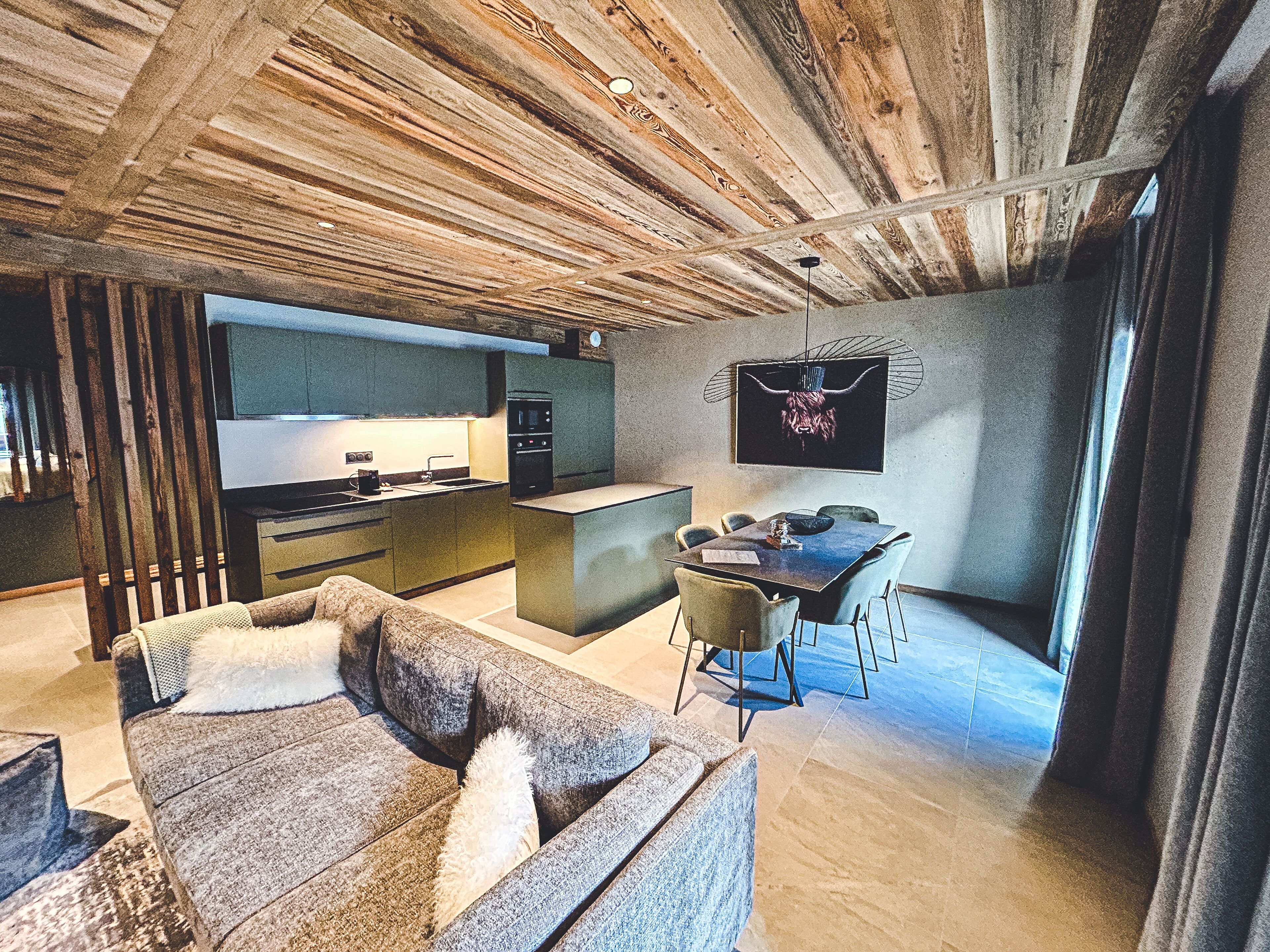 Ein modernes Wohnzimmer mit Sofa, Essbereich mit Tisch und Stühlen, Küchenzeile mit Herd und Spüle sowie einer großen Holzzielung.