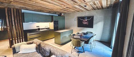 Ein modernes Wohnzimmer mit Sofa, Essbereich mit Tisch und Stühlen, Küchenzeile mit Herd und Spüle sowie einer großen Holzzielung.
