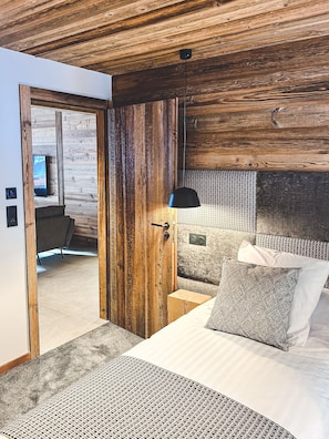 A modern bedroom with a wooden ceiling, a bed with patterned pillows, a bedside table, and a television in the adjacent room. - Hameau des Prodains - Résidence (Morzine)