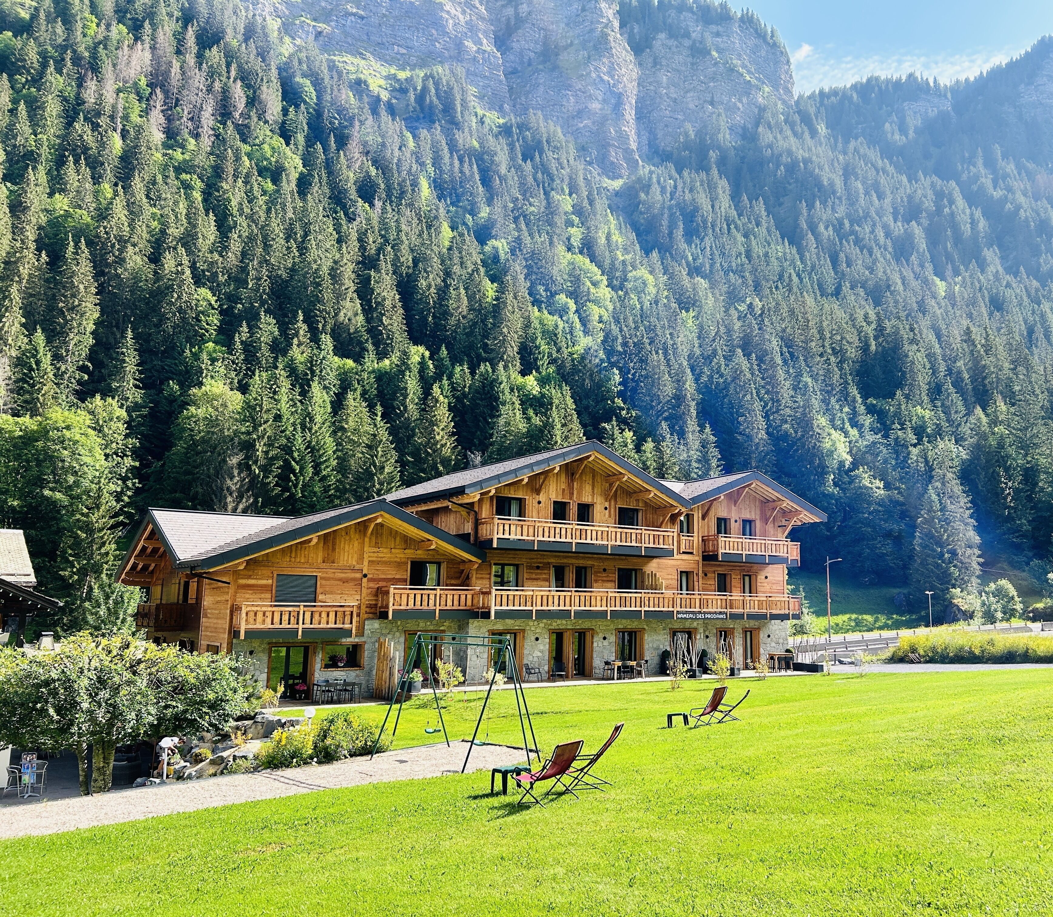 Un chalet en bois doté d’un balcon, entouré d’une pelouse bien entretenue et adossé à une montagne boisée.