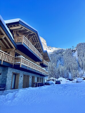 A chalet-style building with wooden balconies and stone walls, surrounded by snow-covered trees and a mountain backdrop. - Hameau des Prodains - Résidence (Morzine)