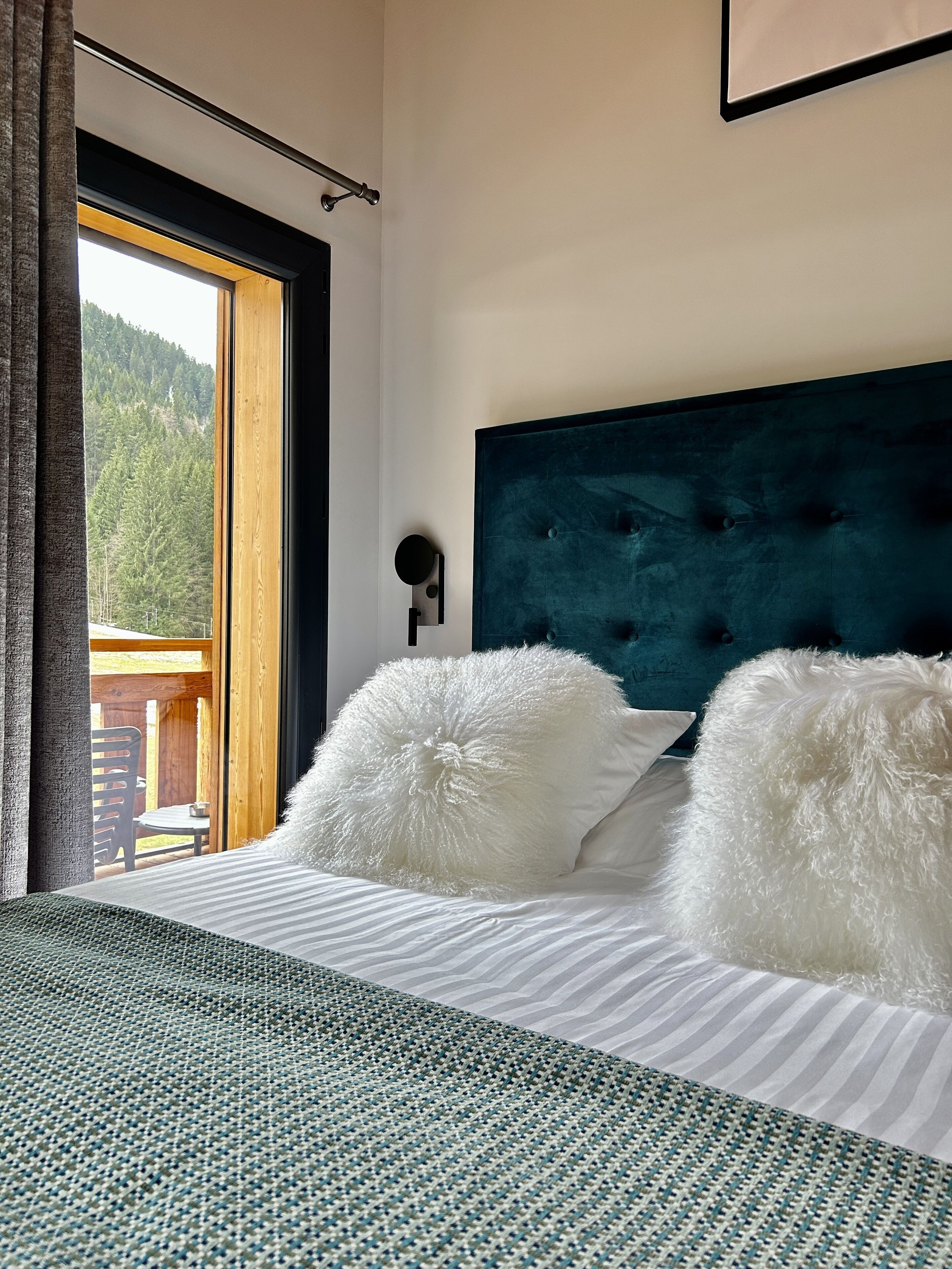 A bedroom with a bed, two white fluffy pillows, a dark headboard, and a view of a wooden deck and forest through a large window.