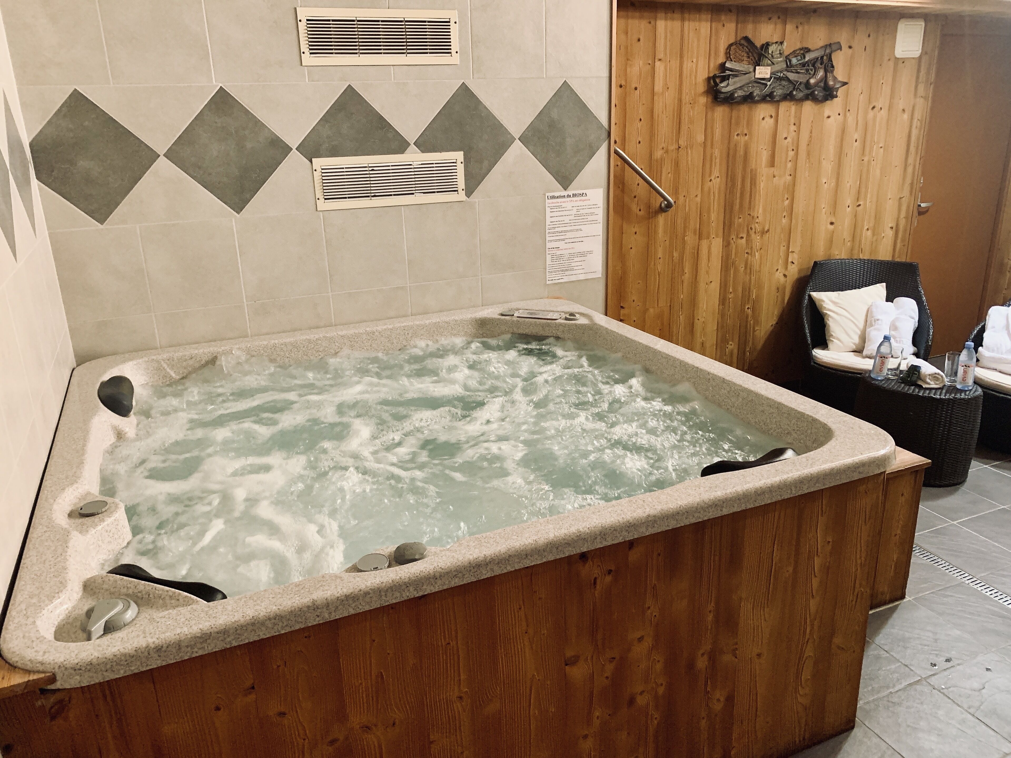 A hot tub with a wooden surround, a wooden door with a towel rack, and a chair with towels and bottles.