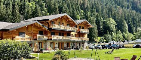 A wooden lodge with multiple balconies, a grassy lawn, wicker furniture, and a mountainous backdrop.