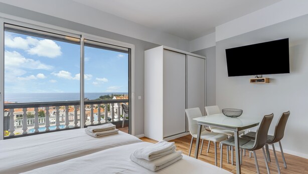 A modern hotel room with two beds, a dining table with chairs, a TV mounted on the wall, and a balcony with a view of the sea and buildings. - Villa Winter Apt. 2 (Funchal)