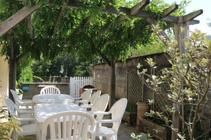 A covered outdoor patio with a table and chairs, surrounded by trees and a white fence.