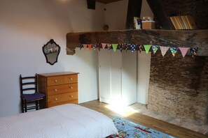 A bedroom with a wooden dresser, a chair, a bed with a checkered bedspread, a mirror, and a wall with a wooden beam and a colorful bunting decoration.