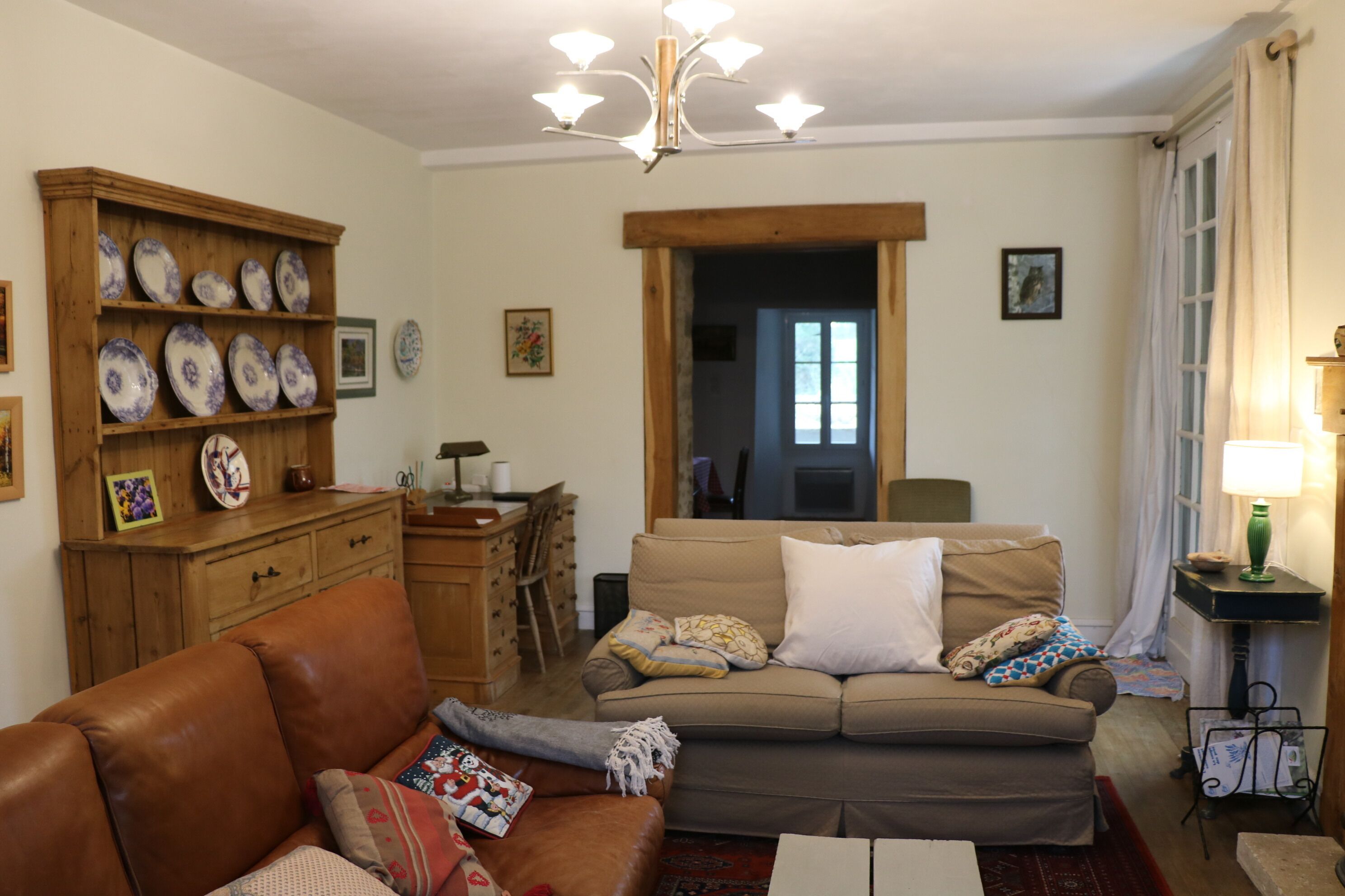 Un salon avec un canapé en cuir marron, un canapé beige et un meuble en bois orné d’assiettes décoratives. On y trouve un lustre, une lampe et une fenêtre avec des rideaux.