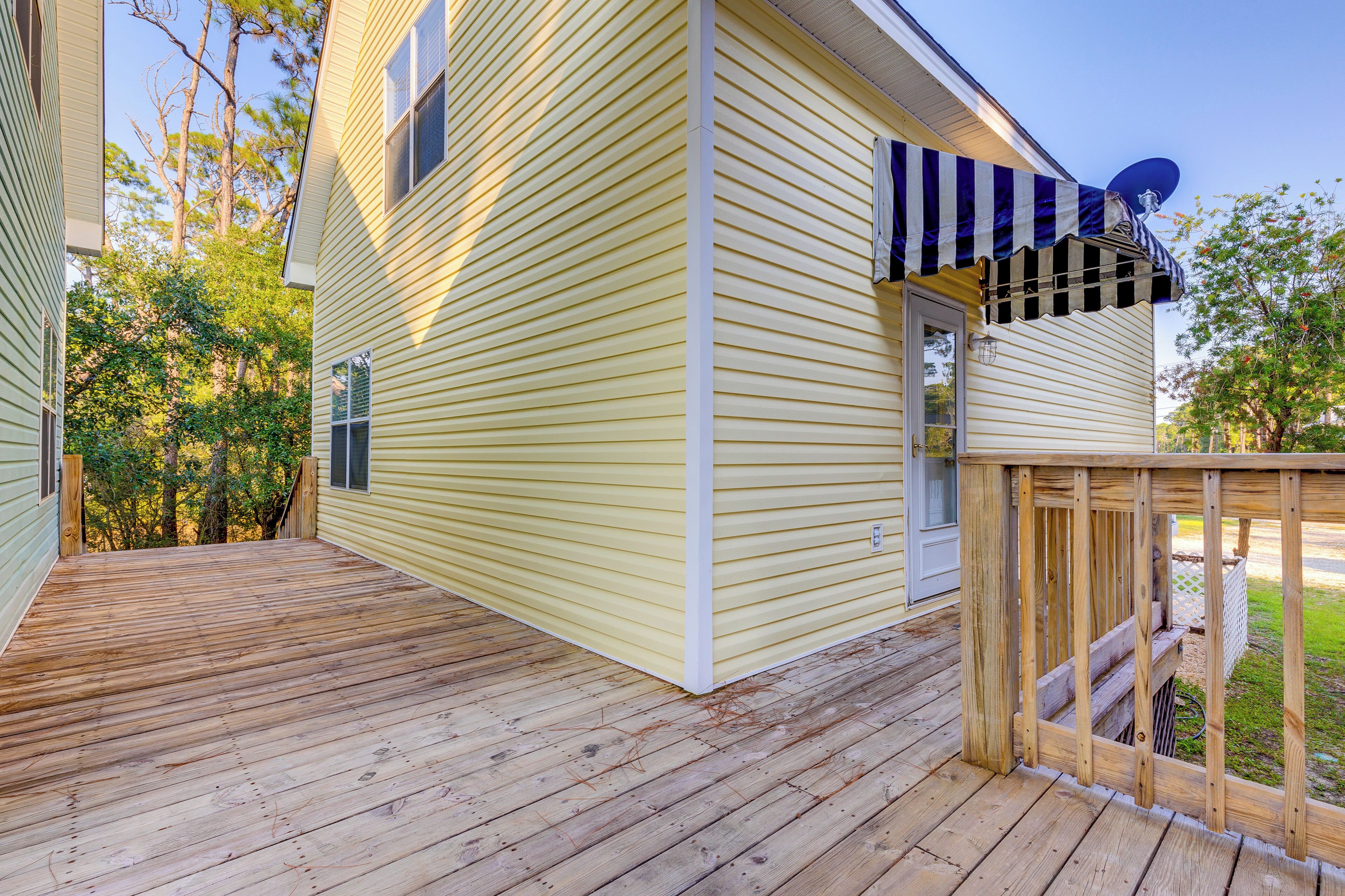 Une terrasse en bois mène à une maison jaune, sur le toit de laquelle on aperçoit une antenne parabolique et qui est dotée d’un auvent à rayures.