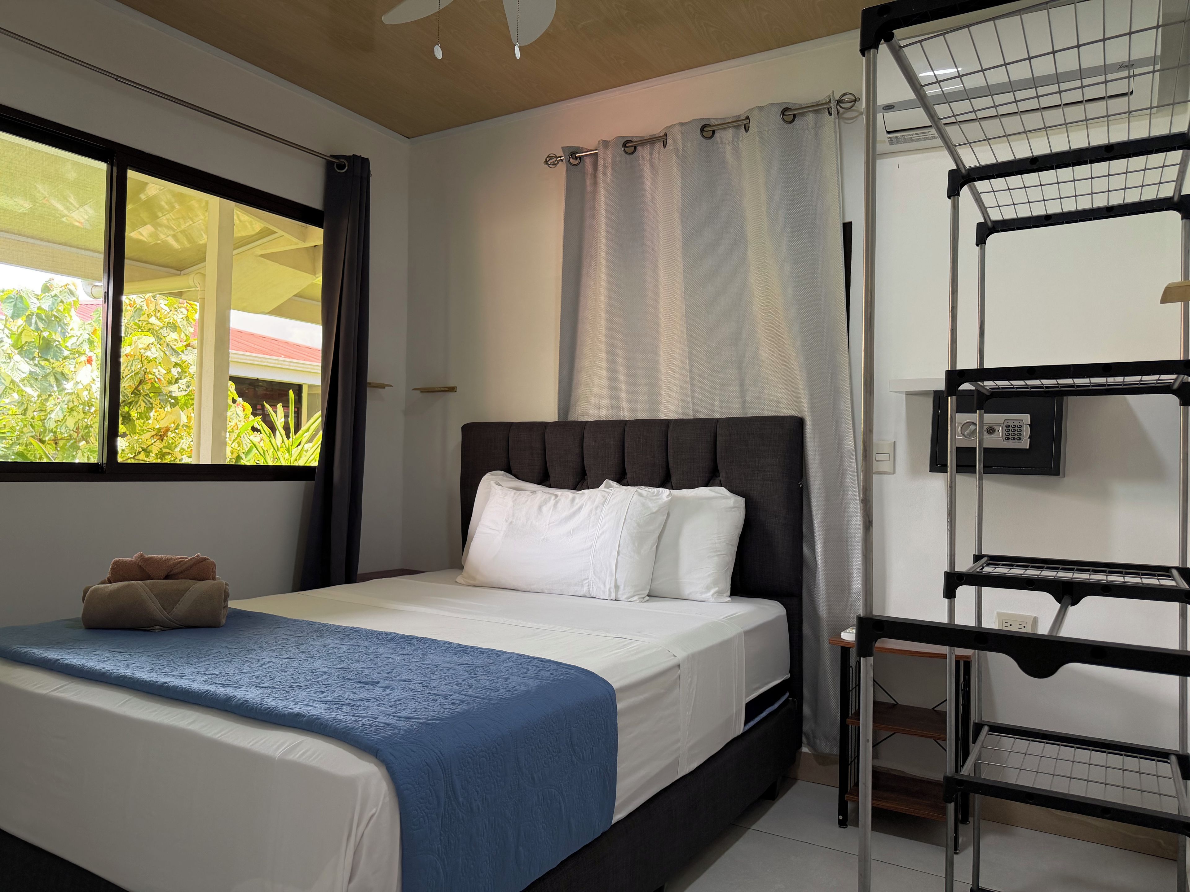 A hotel room with a bed, a window with curtains, a ceiling fan, and a metal shelf unit.