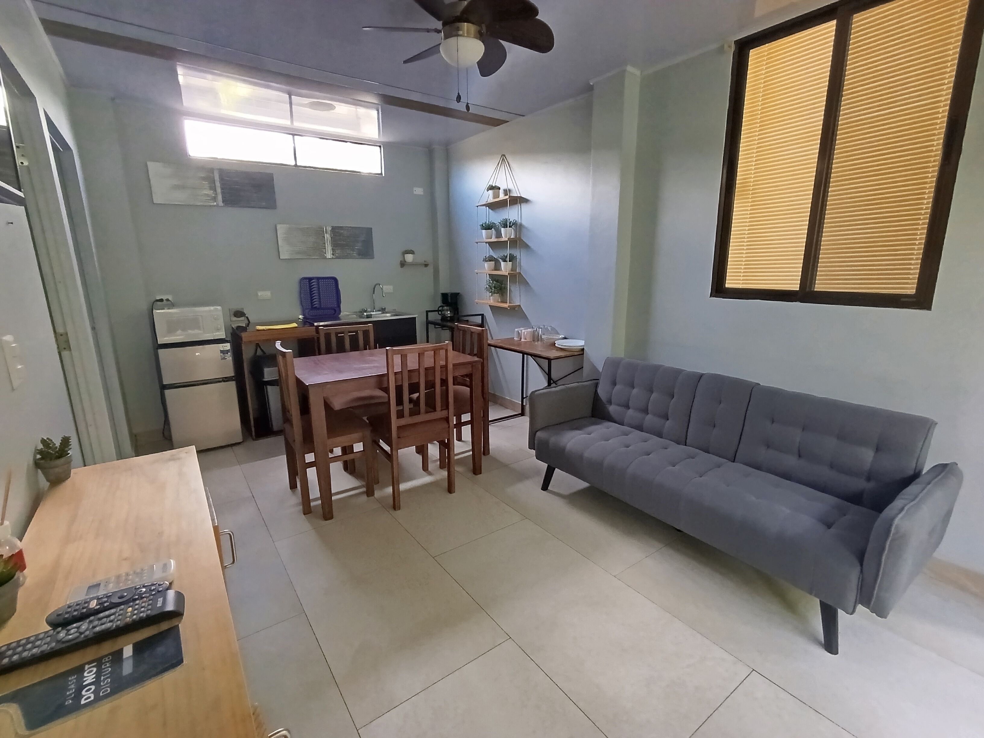 Un espacio habitable compacto con zona de comedor, sofá, ventilador de techo, ventana con persianas y kitchenette.