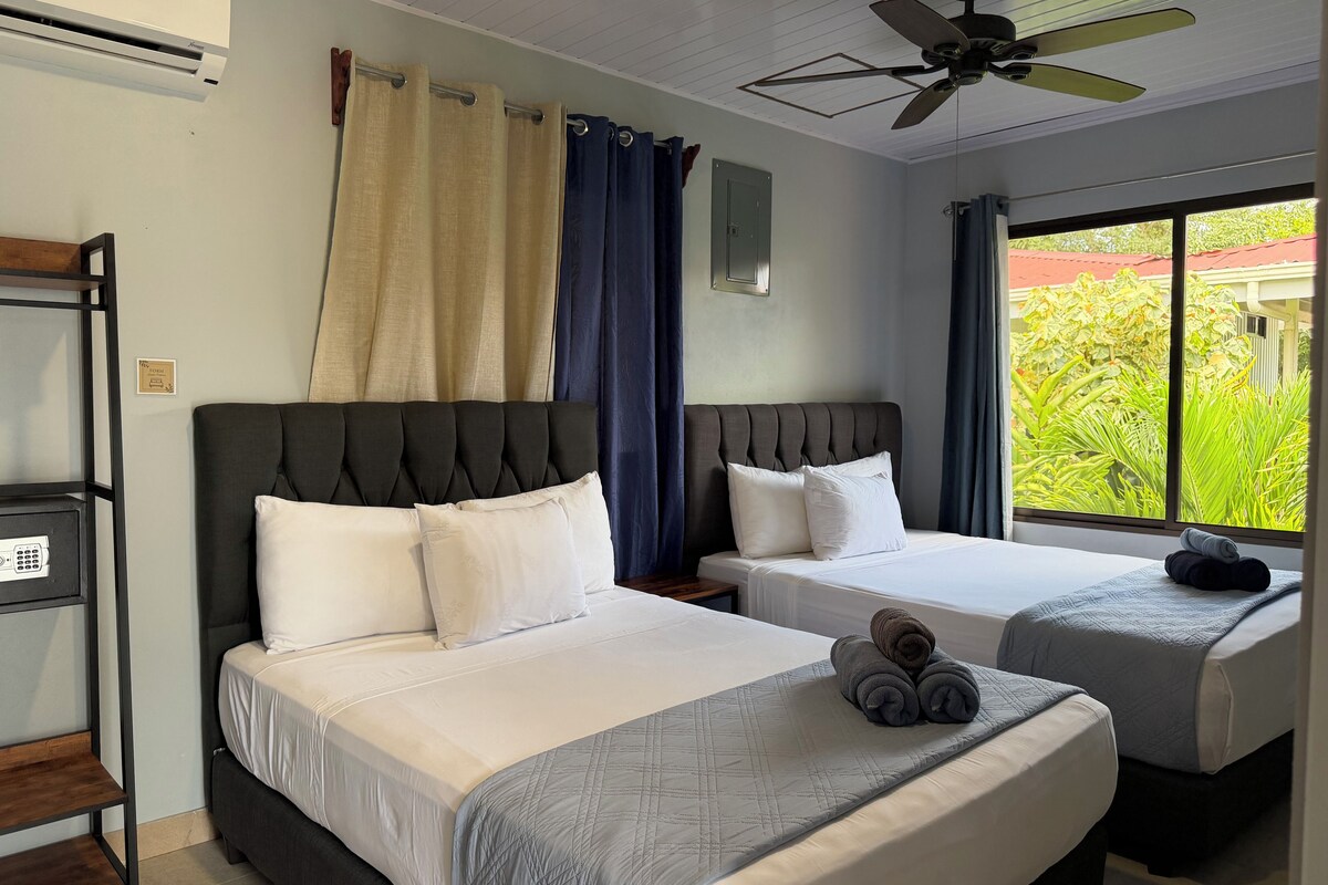 A hotel room with two beds, a ceiling fan, and a view of greenery outside.