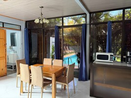Un comedor con mesa y sillas de madera, una cocina con microondas y vistas a una piscina a través de amplios ventanales.