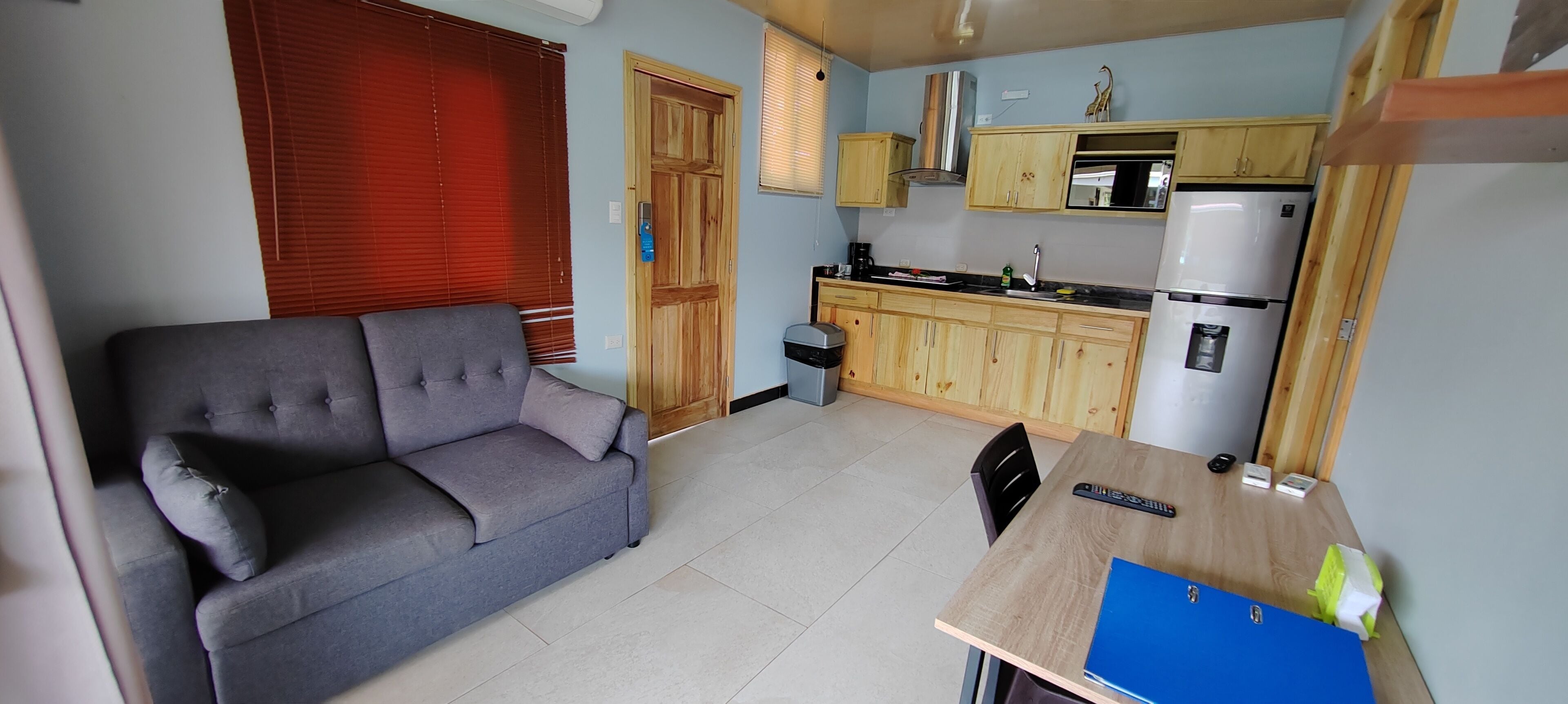 A compact living space with a grey sofa, a wooden dining table with a blue folder, a microwave, and a refrigerator.