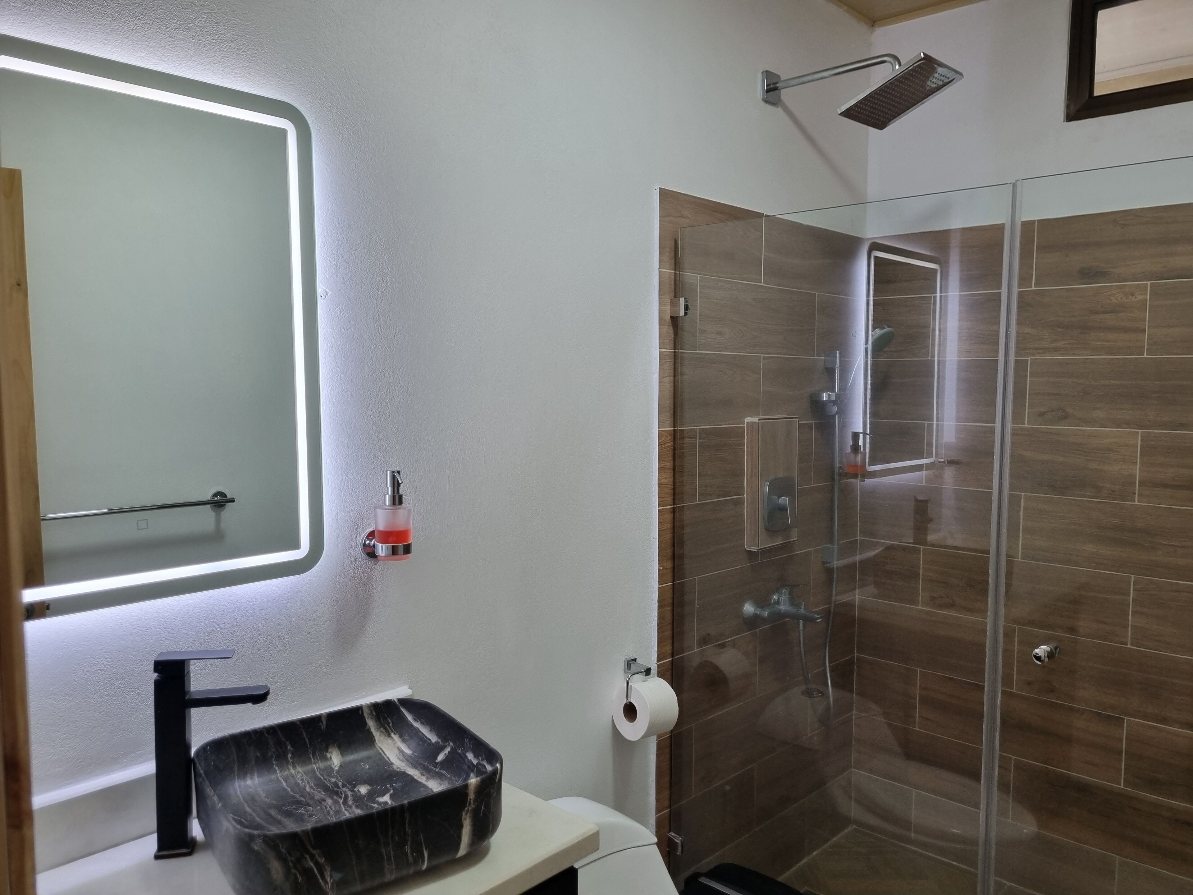 Un baño con ducha acristalada, lavamanos de mármol negro y espejo con luz integrada.