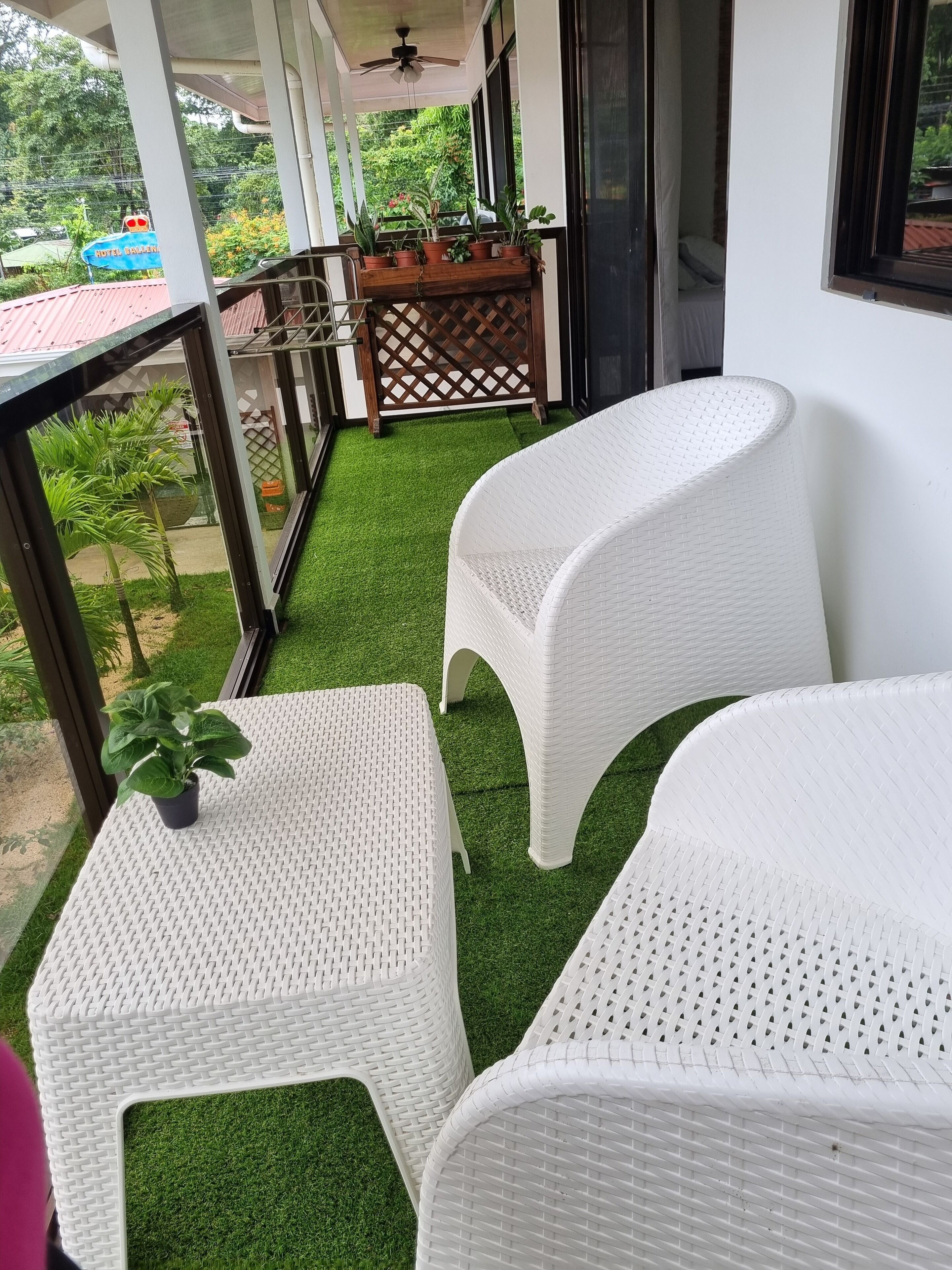 Un balcon doté de meubles en osier blanc et d’un revêtement de gazon synthétique, donnant sur un jardin agrémenté de plantes en pot et d’une enseigne indiquant « Hôtel Bravura ».