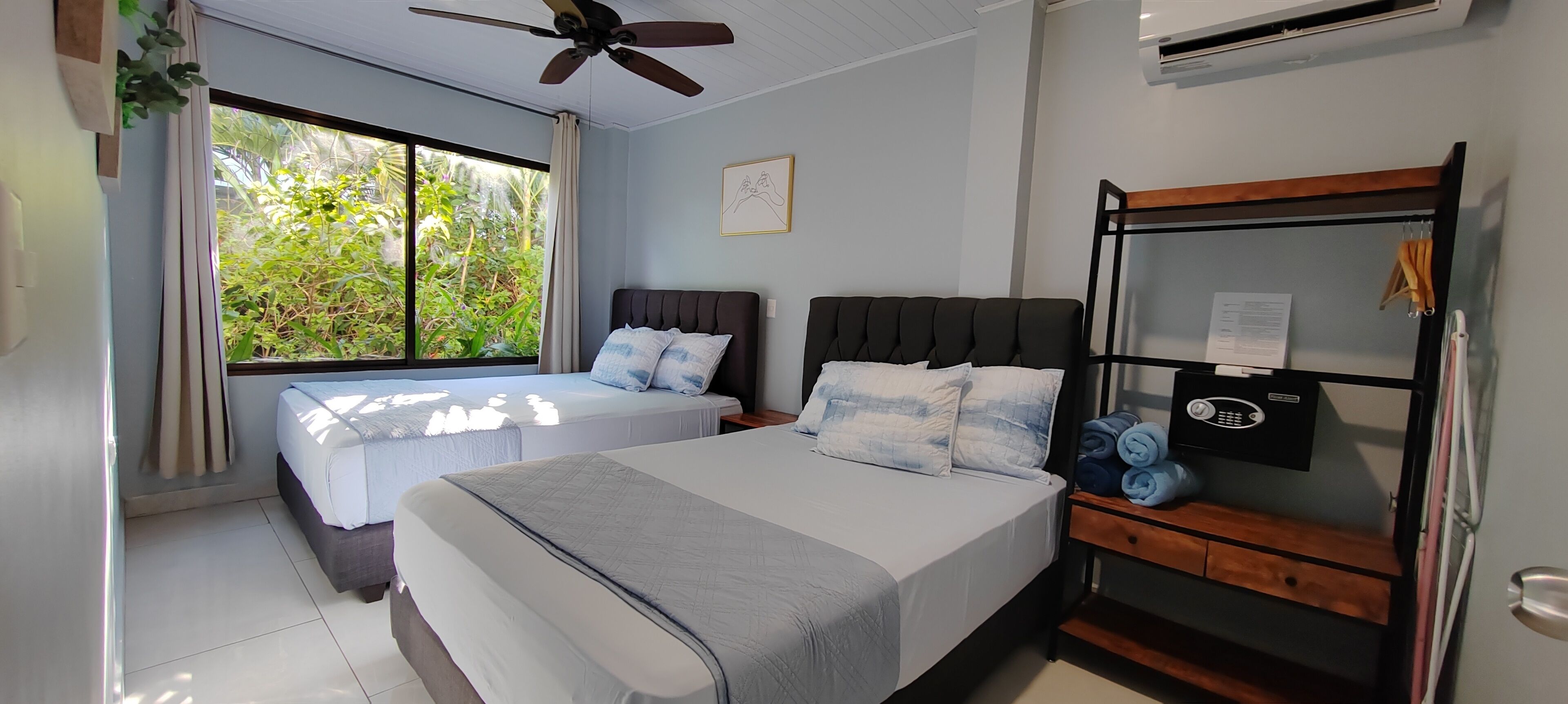 A bedroom with two beds, a ceiling fan, a window with a view of greenery, and a bedside table with a phone and towels.