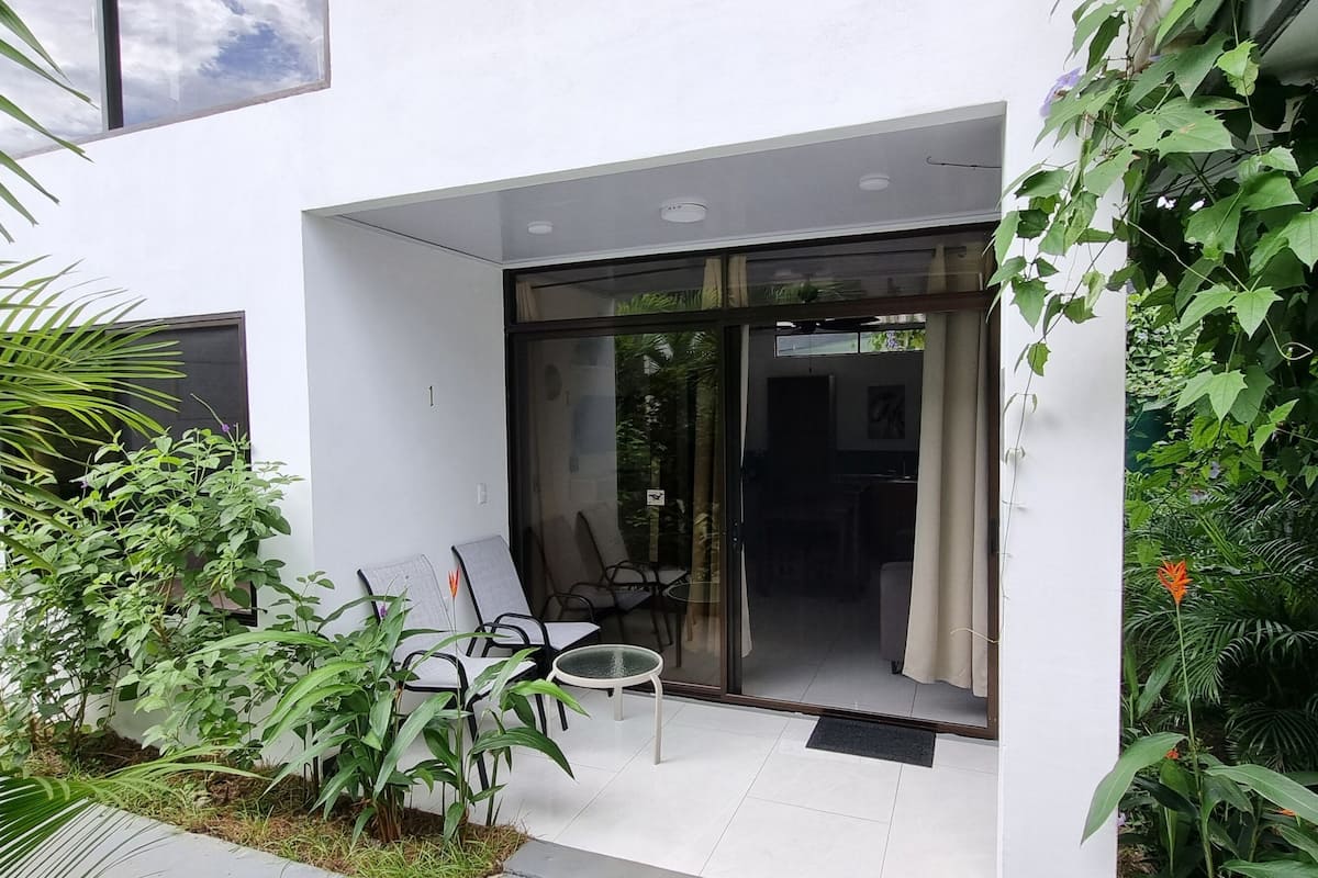 A modern outdoor patio with a glass door leading to a room, surrounded by lush greenery and a small seating area with a table and chairs.