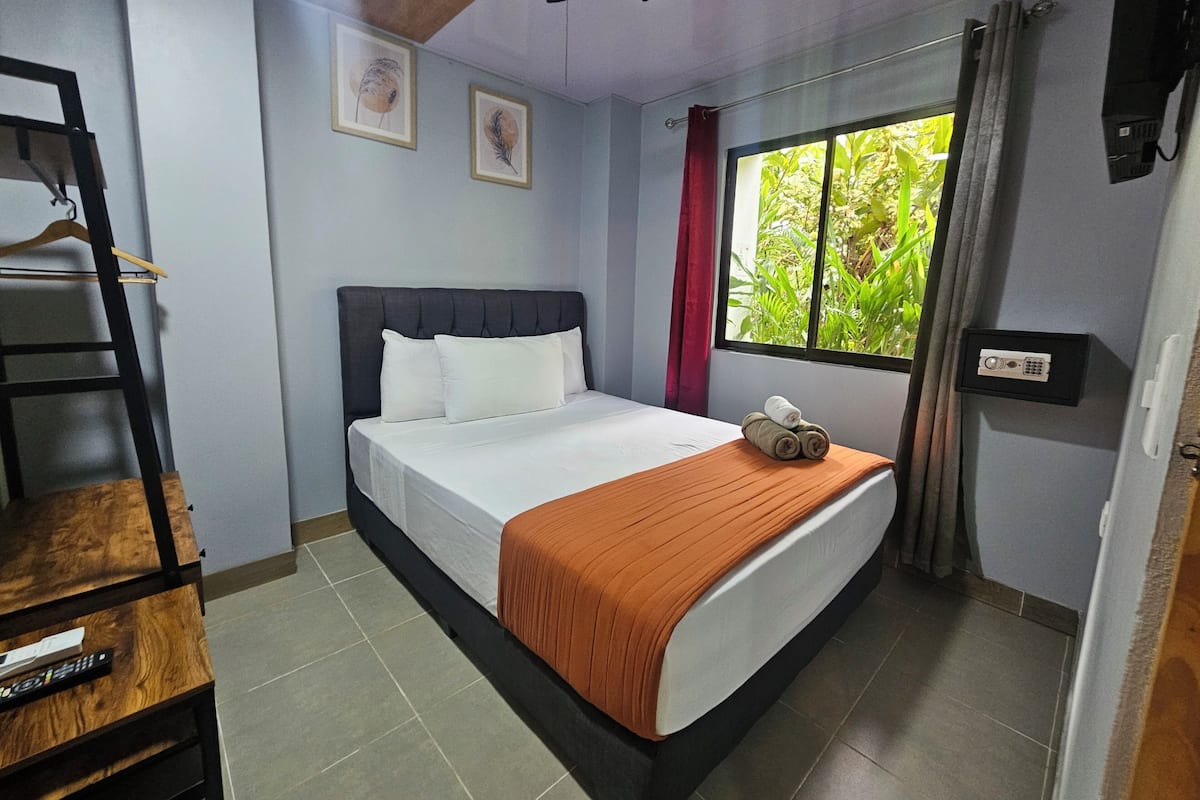 A bedroom with a bed, a ceiling fan, a window with curtains, a bedside table with a remote control, and a wall-mounted speaker.