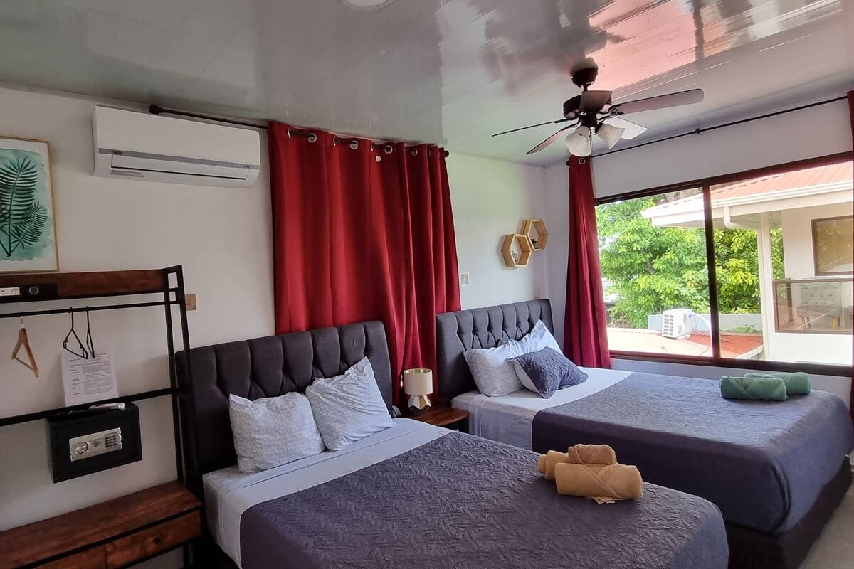A hotel room with two beds, a ceiling fan, an air conditioning unit, and a window with red curtains.