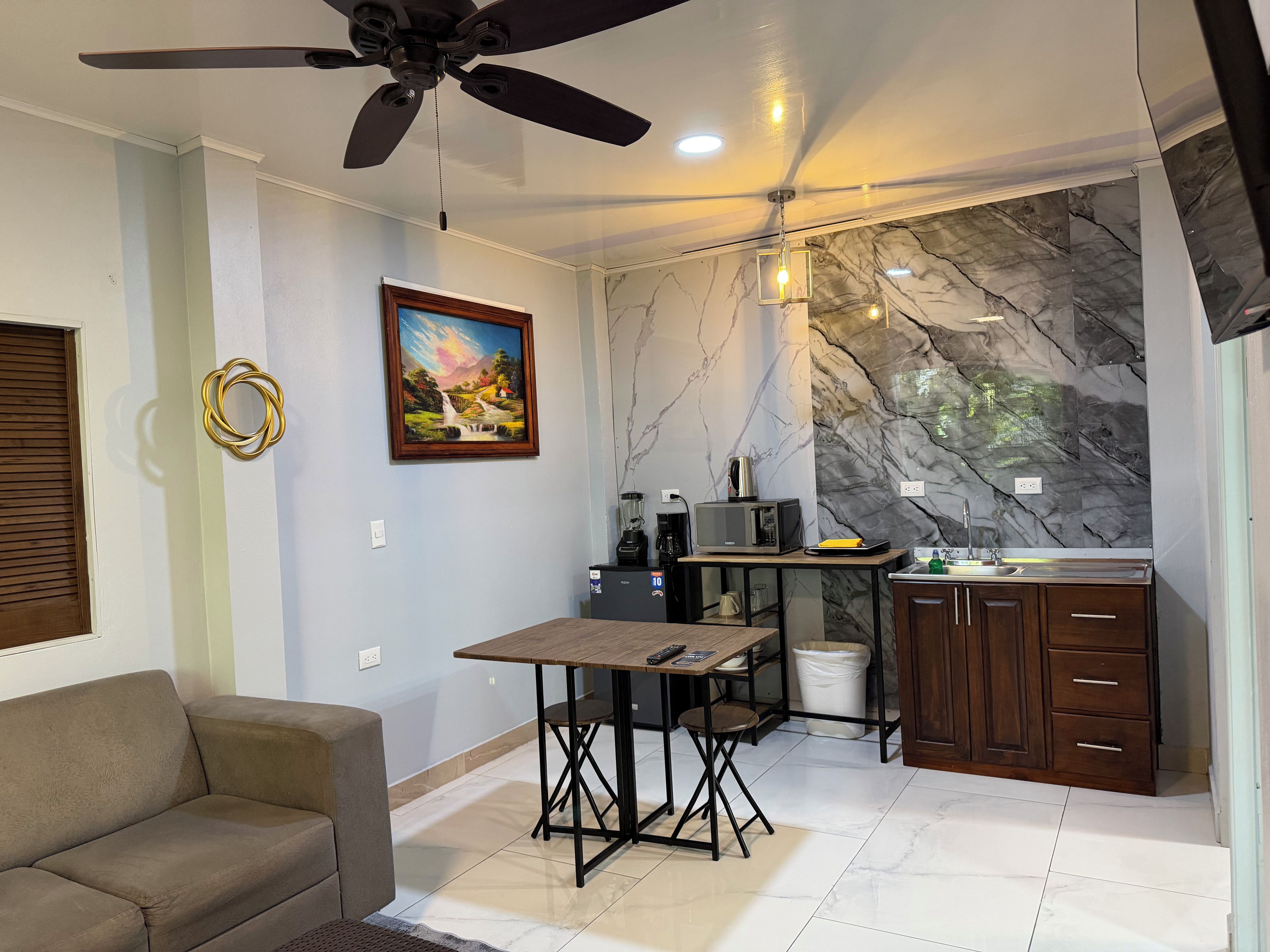 A compact living space with a ceiling fan, a dining table, a sofa, a kitchen area with a marble backsplash, and a painting on the wall.