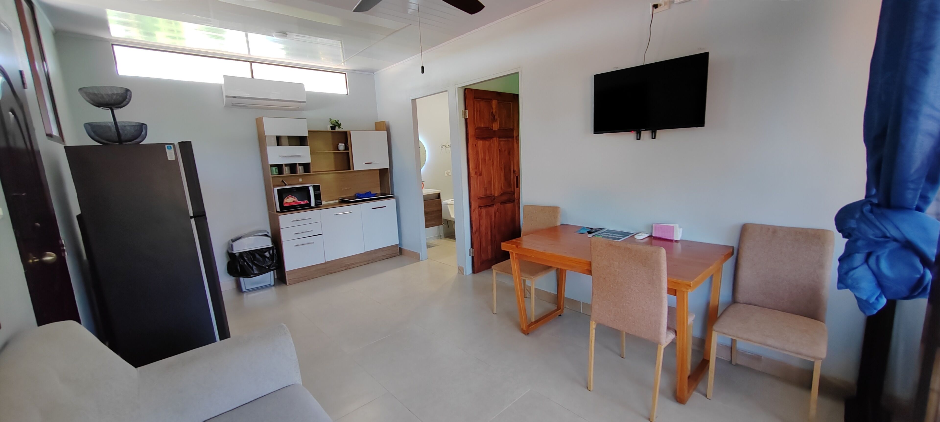 A modern kitchen with a refrigerator, microwave, and wooden cabinets. There is a dining table with chairs and a TV mounted on the wall.