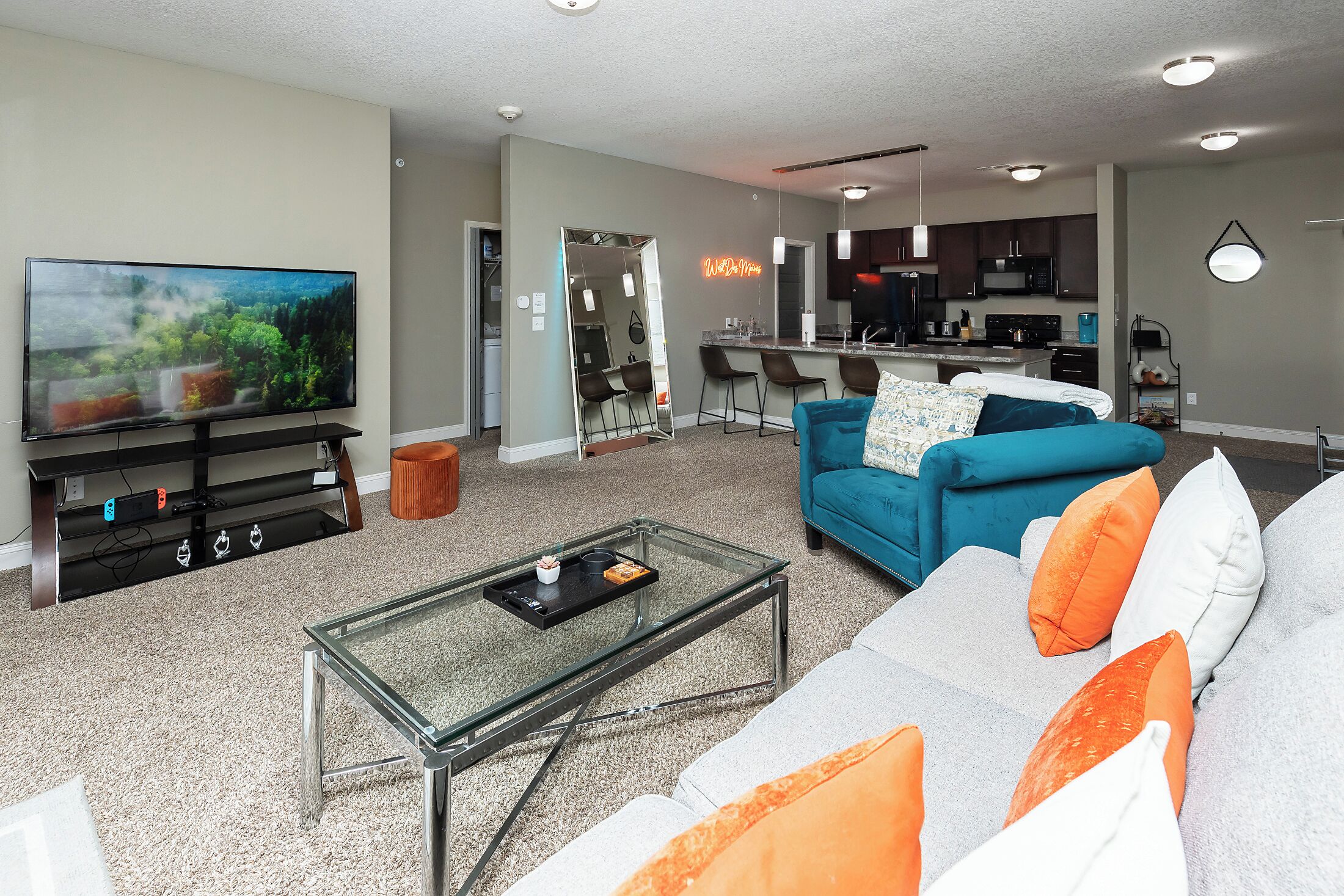 A modern living room with a flat-screen TV, a glass coffee table, and a sofa with orange and white cushions.