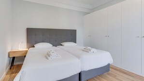 A modern hotel room with two beds, a grey headboard, white bedding, a bedside table with a lamp, and a wardrobe. - Villa Winter Apt. 3 (Funchal)