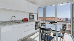 A modern kitchen with white cabinets, a granite countertop, and a red toaster. There is a dining table with chairs and a large window offering a view of the city. - Villa Winter Apt. 3 (Funchal)