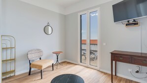 A room with a wooden floor, a round table, a chair, a small stool, a desk, a television mounted on the wall, and a sliding glass door leading to an outdoor area. - Villa Winter Apt. 3 (Funchal)