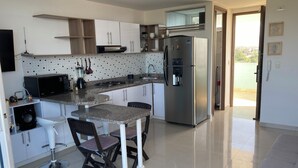 A modern kitchen with white cabinets, a stainless steel refrigerator, a microwave, and a gas stove. There are two bar stools and a dining table.