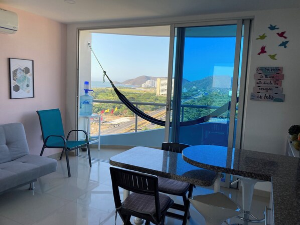 Una sala moderna con un sofá blanco, una mesa de comedor con sillas, una hamaca y un ventanal con vistas a un paisaje urbano y colinas.