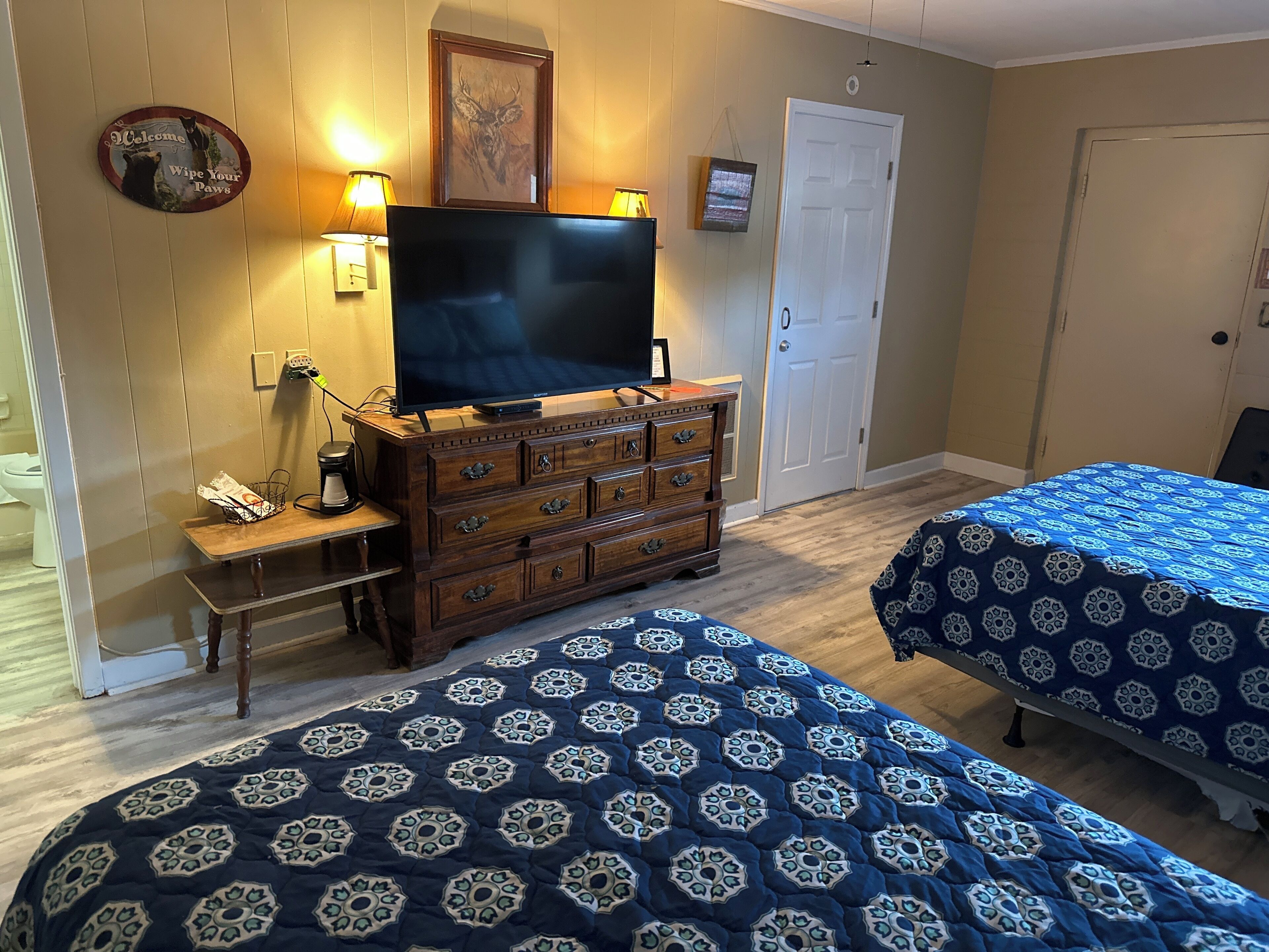 Habitación con dos camas, un televisor sobre un aparador, una cafetera y un cartel de bienvenida.