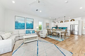 A modern living room with a sectional sofa, a dining area with a wooden table and chairs, and a kitchen with white cabinets and stainless steel appliances.