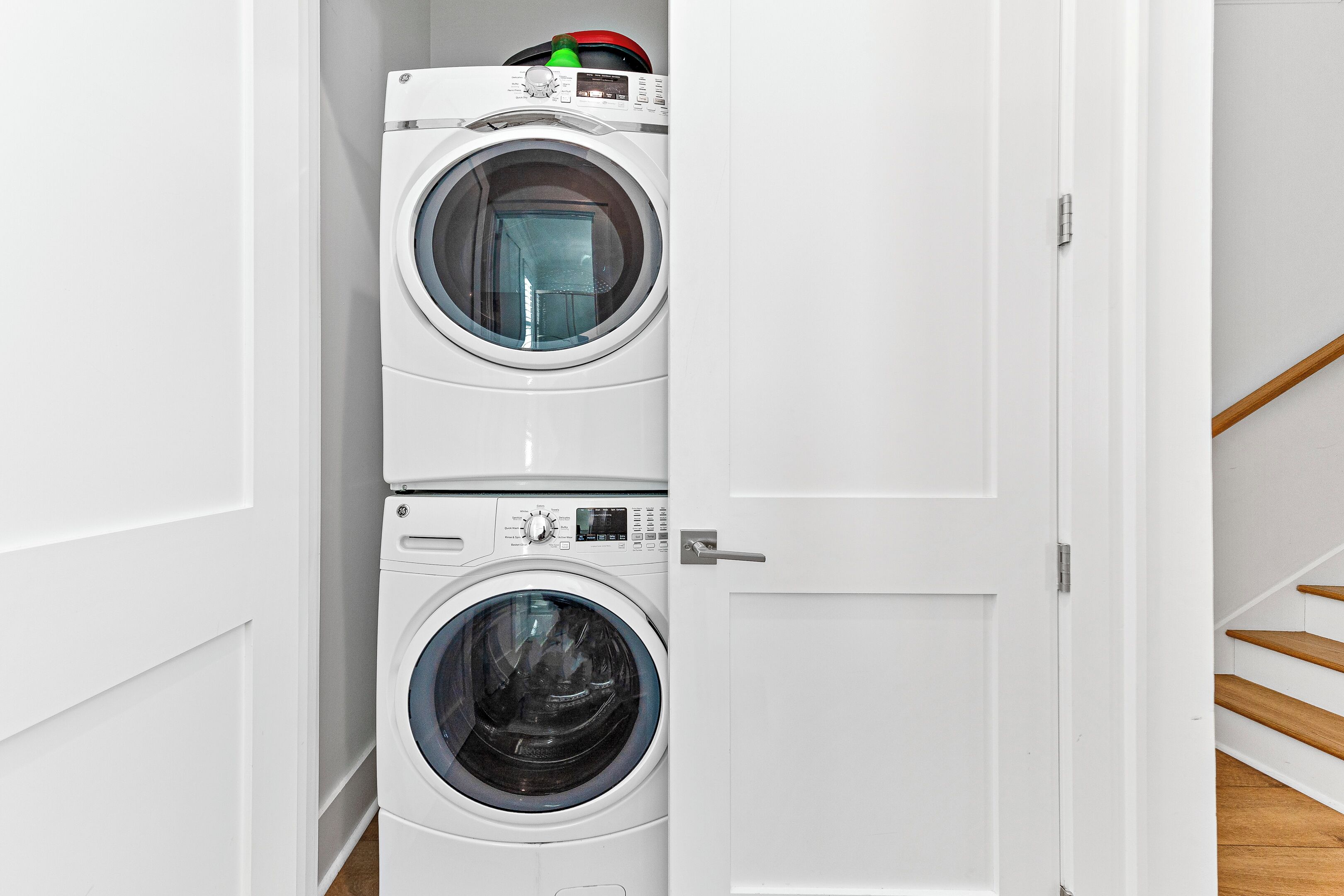 A stacked washer and dryer unit in a hallway with a wooden staircase.