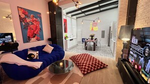 Een moderne woonkamer met een blauwe bank, een ronde salontafel en een geruit kussen. Er hangt een schilderij aan de muur, er staat een televisie en een lamp.