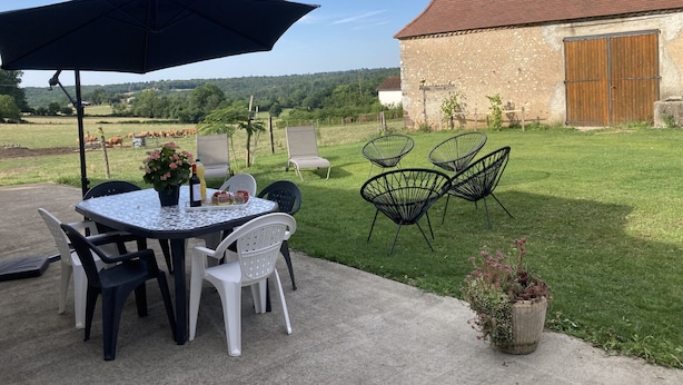 Une terrasse extérieure avec une table et des chaises, un grand parasol, et une vue sur un bâtiment en pierre et des champs.