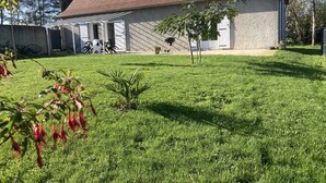Un bâtiment avec un toit recouvert de tuiles rouges, un patio abrité avec des chaises, et un petit arbre au premier plan.