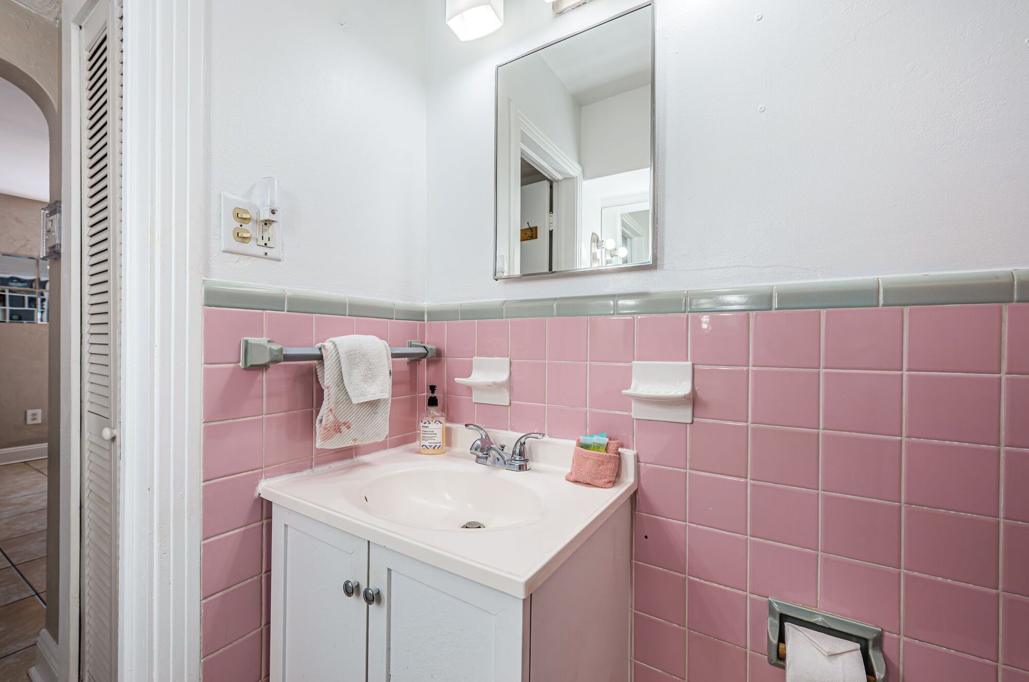 A bathroom with a white sink, pink tiles, a towel rack, a mirror, and a soap dispenser.