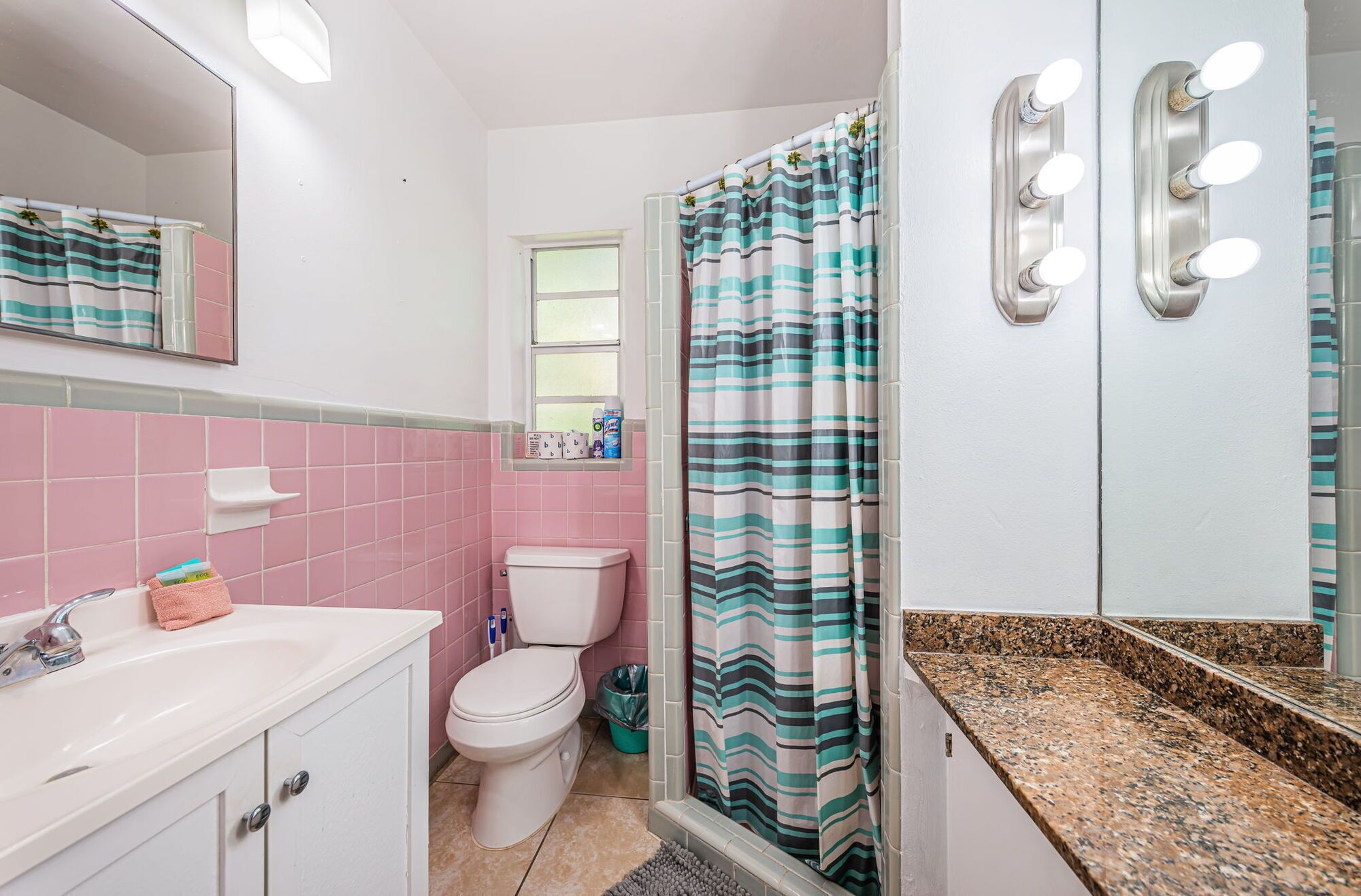 Ein Badezimmer mit rosafarben gefliesten Wänden, einem weißen Waschbecken mit Spiegel, einer Toilette und einer Dusche mit gestreifter Duschvorhang.